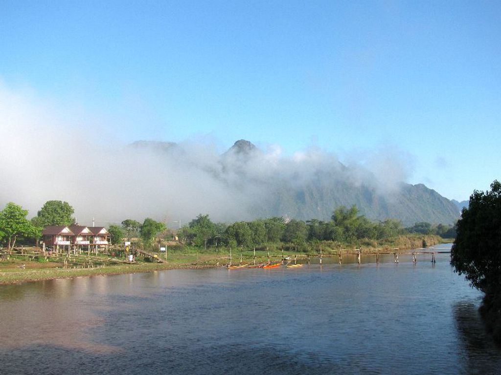 Rivierlandschap in Laos met houten brug, dorp en bergen in de mist langs de Mekong.