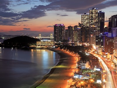Avondzicht op skyline van Busan met strand, verlichte brug en hoogbouw langs de kust.