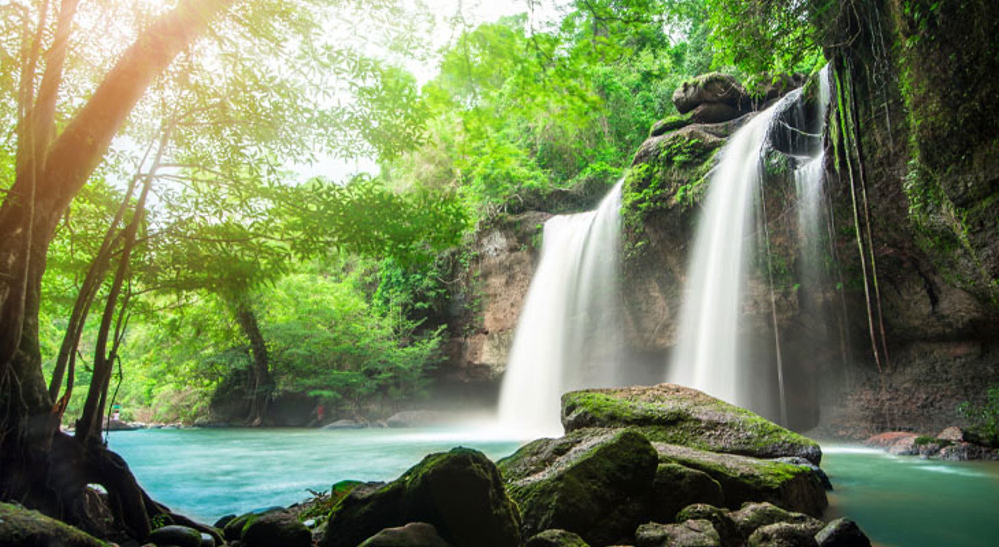 Waterval in een groene jungle in Thailand met turquoise water en rotsen op de voorgrond.