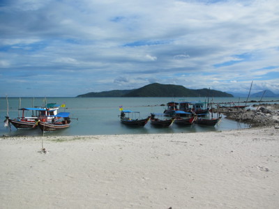 Longtailboten voor anker bij een wit zandstrand in Zuid-Thailand, met een groen eiland op de achtergrond.