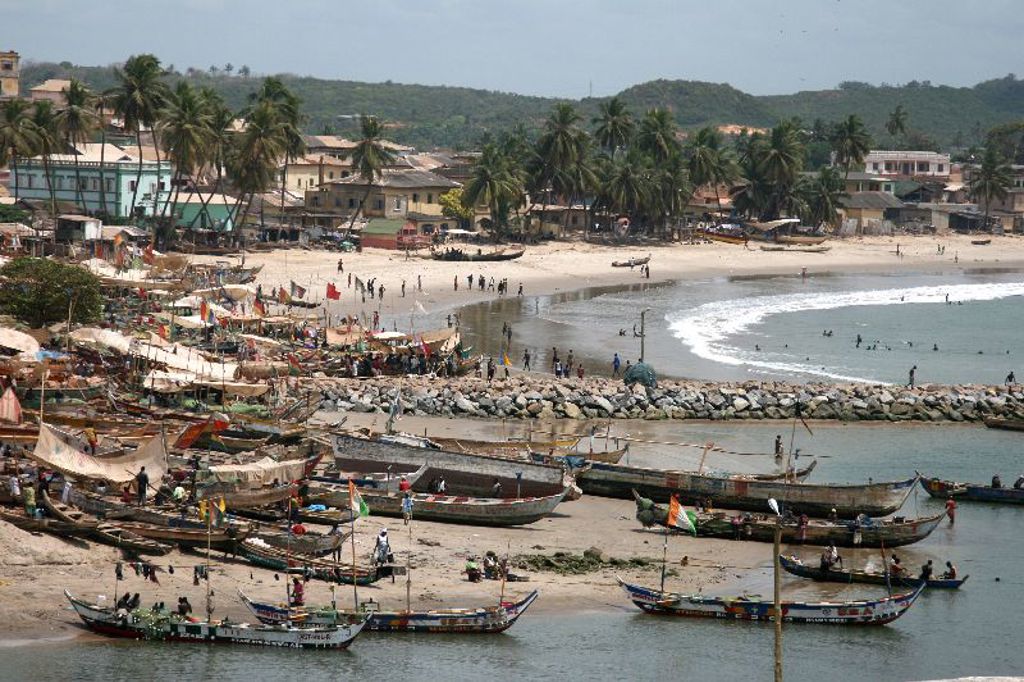 Vissersboten bij een druk strand met palmbomen en huizen aan de kust in Ghana.