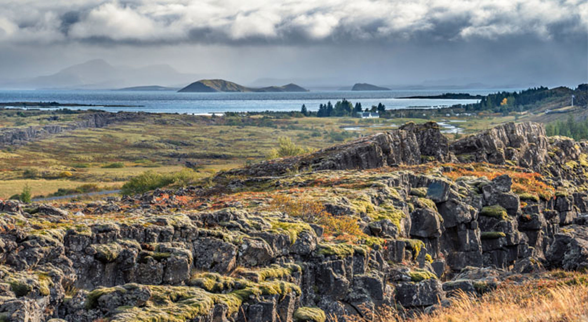 Uitzicht over het Þingvellir Nationaal Park in IJsland met rotskloven, mos en een meer onder bewolkte lucht.