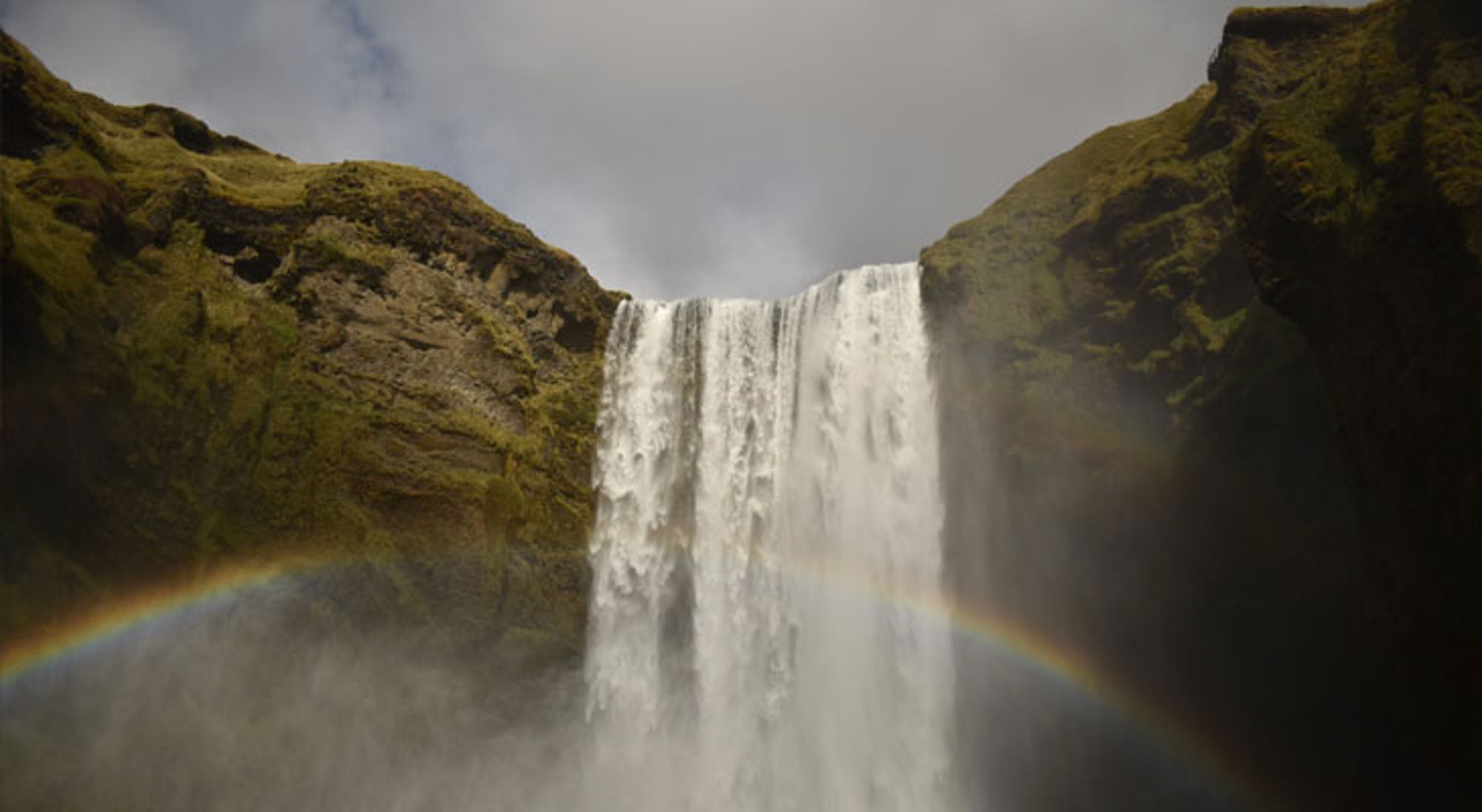 Waterval Skógafoss in IJsland met regenboog in de nevel, omringd door groene rotswanden.
