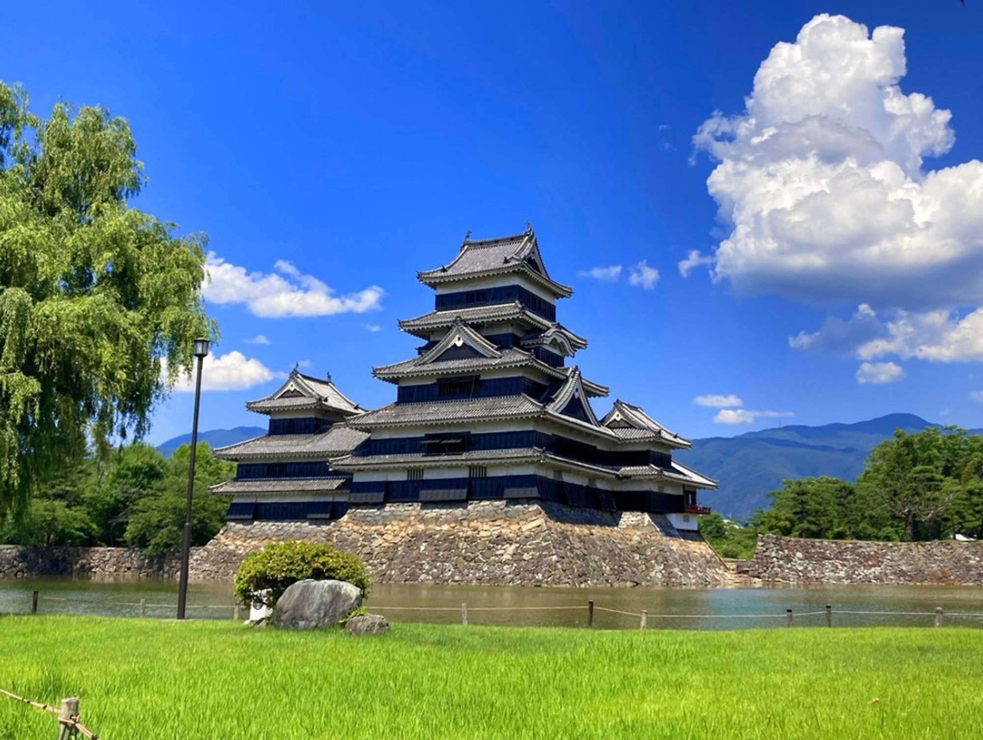 Matsumoto-kasteel in Japan met gracht, groene tuin en bergen onder een blauwe lucht.