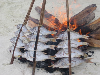 Vis wordt op houten stokken boven een kampvuur op het strand geroosterd.