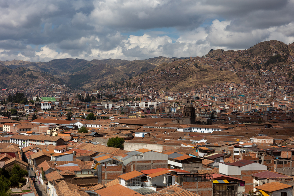 Panorama van Cusco in Peru met rode daken, een kerktoren en bergen onder een bewolkte lucht.
