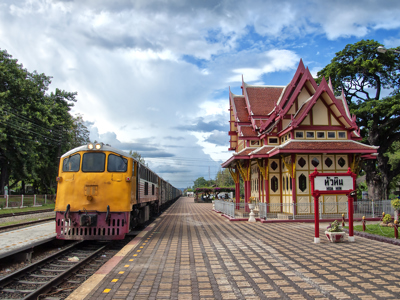 Geel-rode trein bij het station van Hua Hin langs een Thais stationsgebouw met puntdak.
