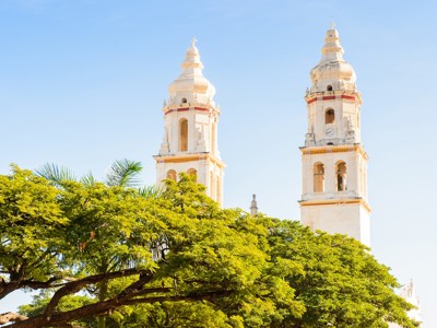 Witte kathedraaltorens boven groene bomen in Campeche op de Route Mexico.