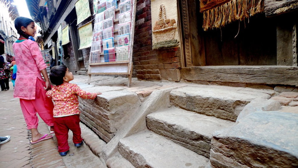 Moeder en kind lopen door een smalle straat met oude stenen trappen en winkeltjes in Nepal.
