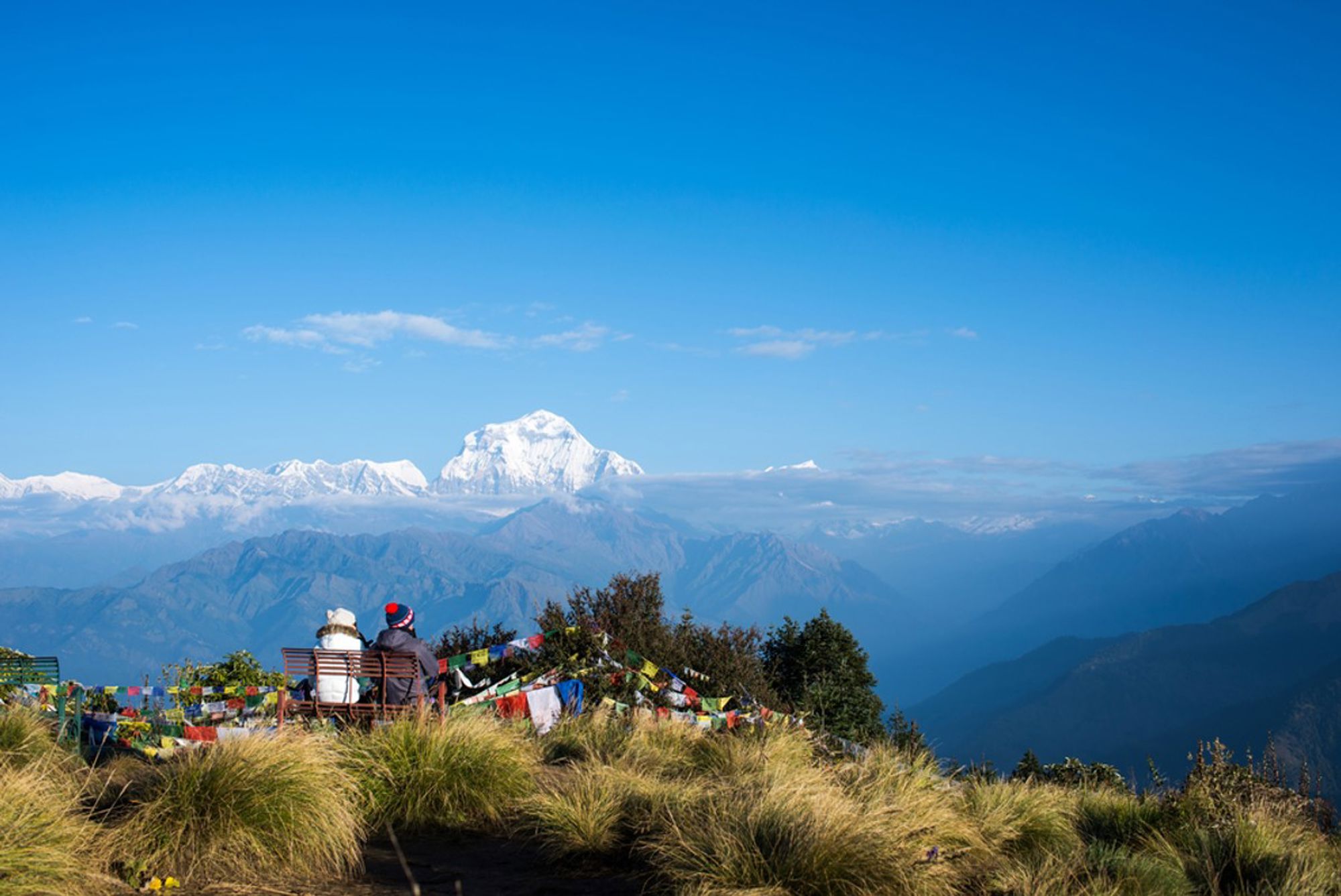 Uitzicht op de besneeuwde Himalaya in Nepal, met twee wandelaars bij gebedsvlaggen op een bergkam.