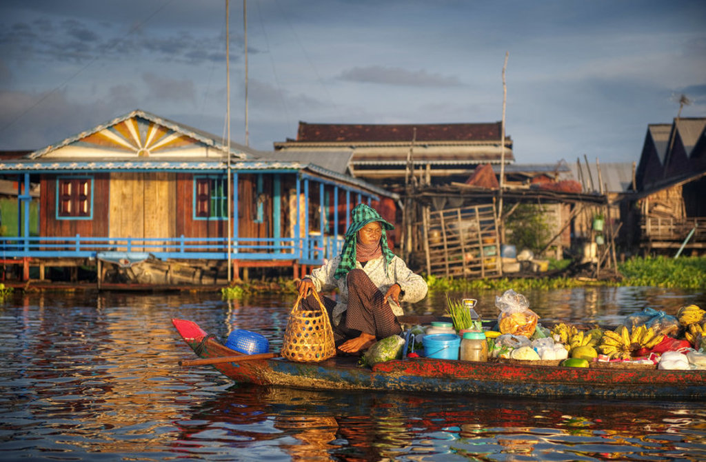 Drijvende markt in Thailand met verkoper in boot vol fruit en groenten voor huizen op palen.