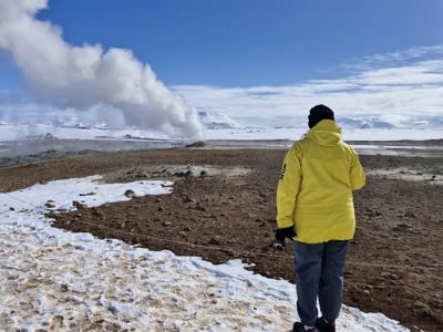 Reiziger bij geothermisch gebied met stoompluim in Noord-IJsland, omringd door sneeuw en bergen.