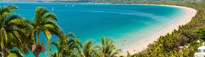 Uitzicht op het strand en de turquoise baai bij Cairns, met palmbomen en bergen op de achtergrond.
