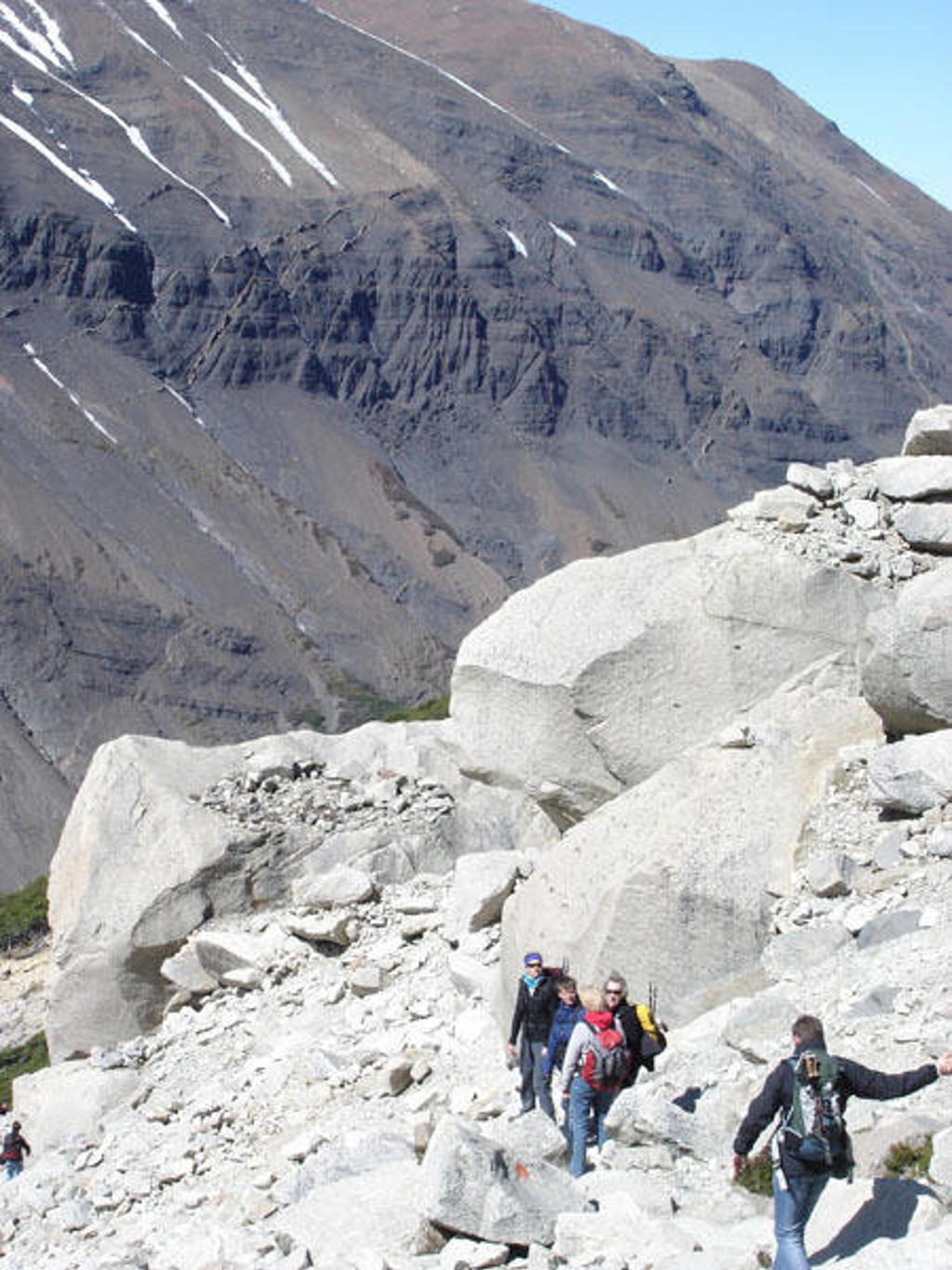 Wandelaars op rotsachtig bergpad in Patagonië, met steile bergwand en sneeuwresten op de achtergrond.