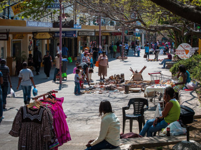 Drukke winkelstraat in Windhoek met marktkraampjes en voetgangers onder schaduwrijke bomen.