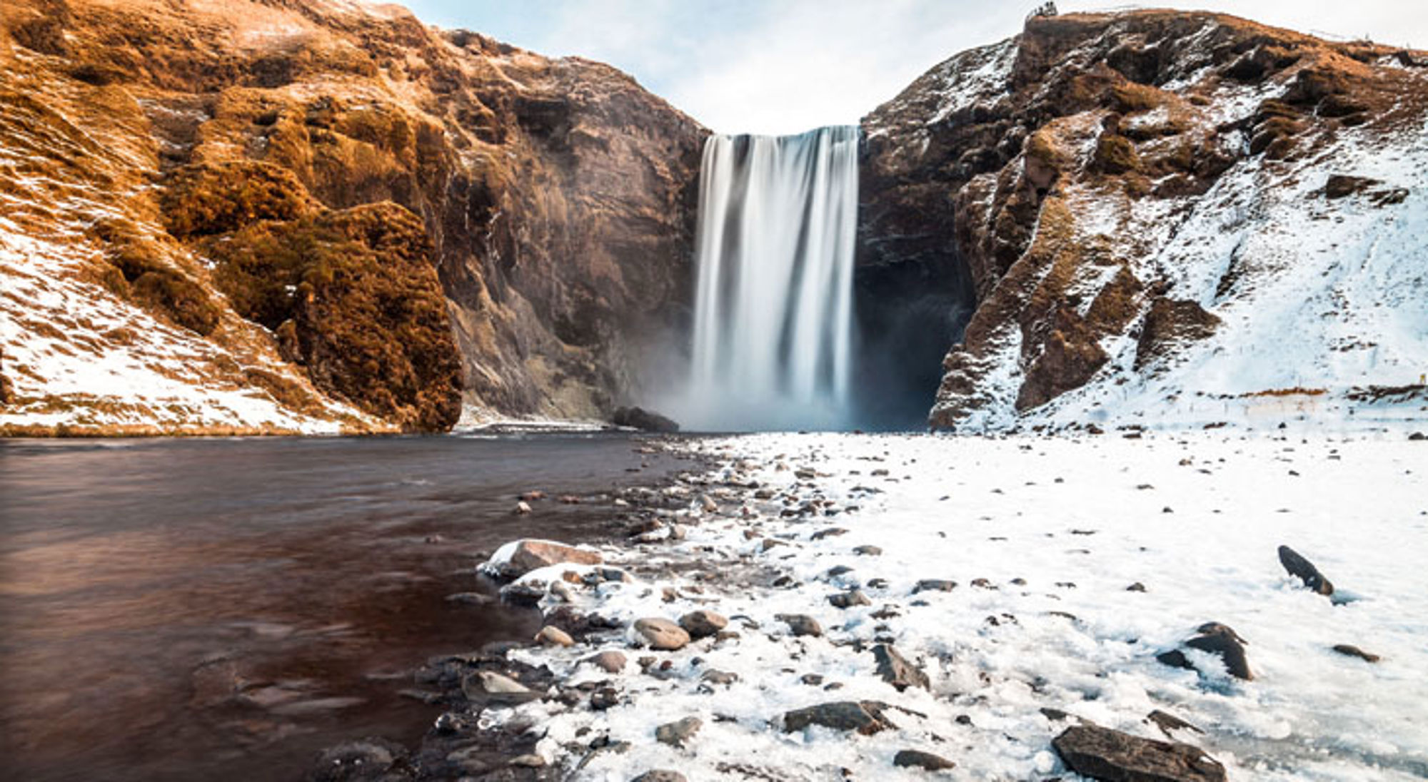Skógafoss-waterval in IJsland met besneeuwde rivierbedding en rotsachtige heuvels.