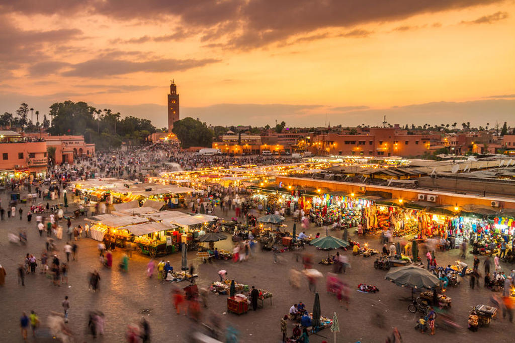 Drukke Djemaa el Fna-plein in Marrakech bij zonsondergang, met marktstalletjes en de Koutoubia-minaret.