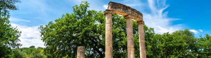 Ruïnes van de tempel van Zeus in Olympia, Griekenland, met zuilen tussen groene bomen onder blauwe lucht.