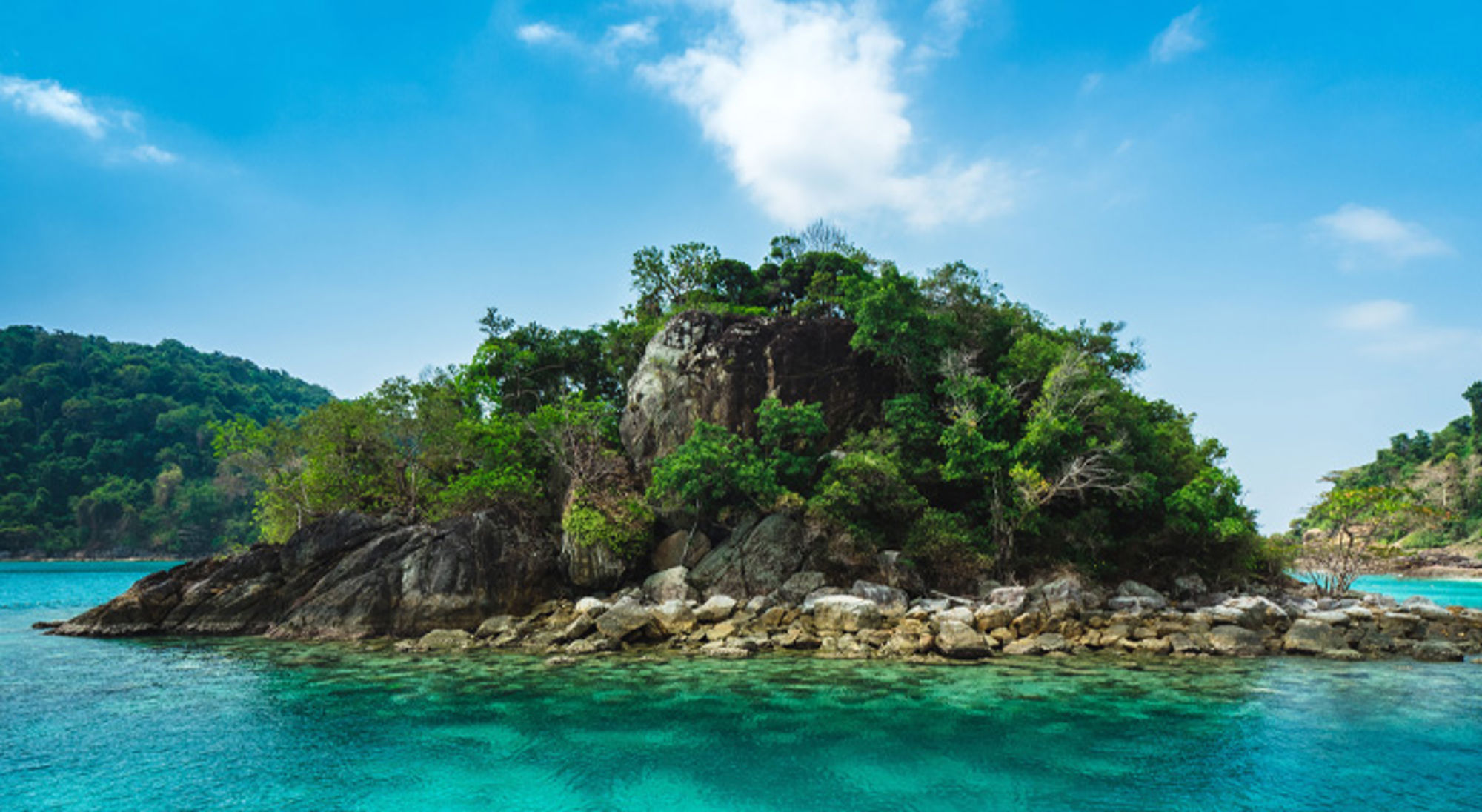 Klein rotsachtig eiland met groene begroeiing in helder turquoise zee in Thailand.
