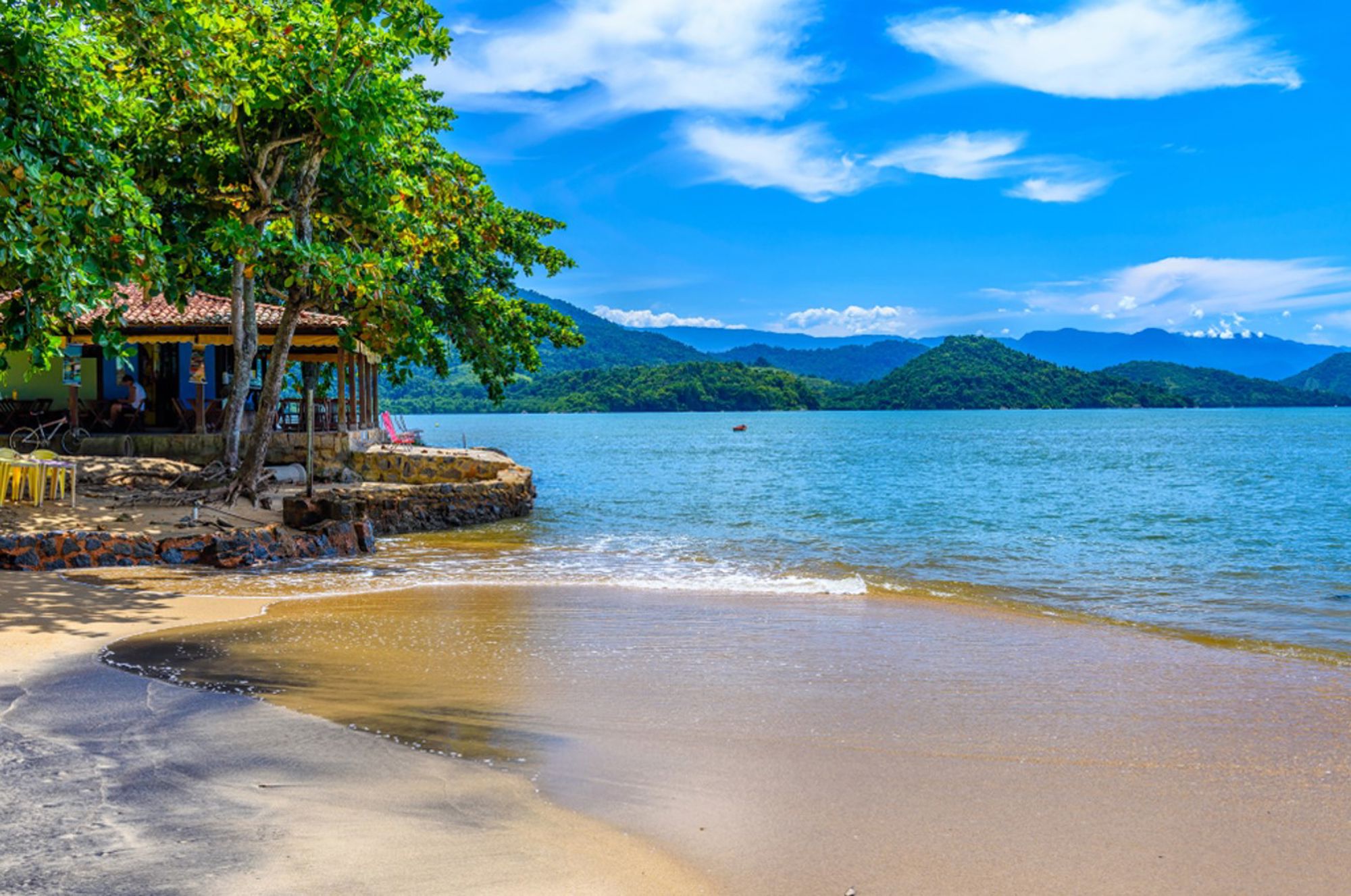 Rustig strand met bomen en strandhuisje aan een baai, met groene heuvels op de achtergrond in Brazilië.