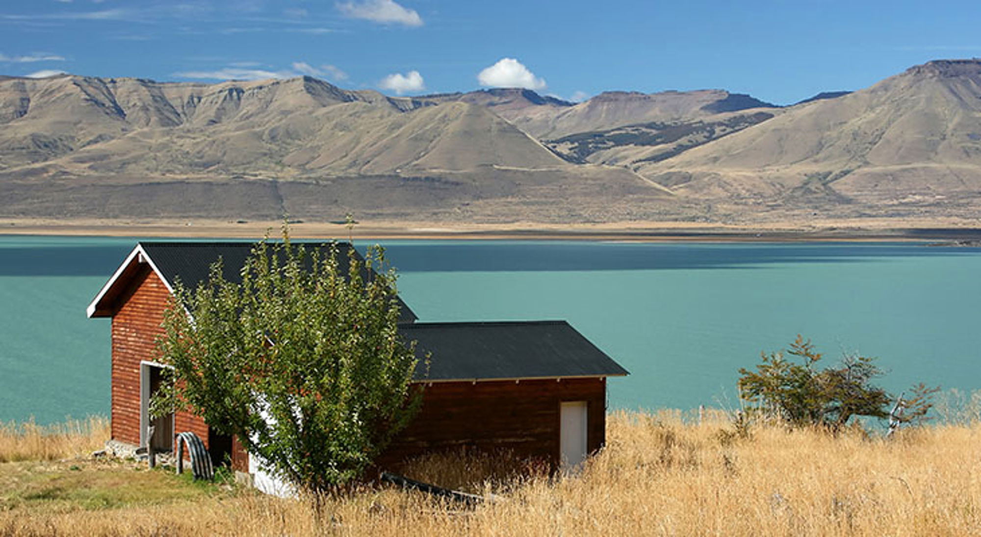 Rood houten huisje aan een turkoois meer met bergen op de achtergrond in Patagonië.