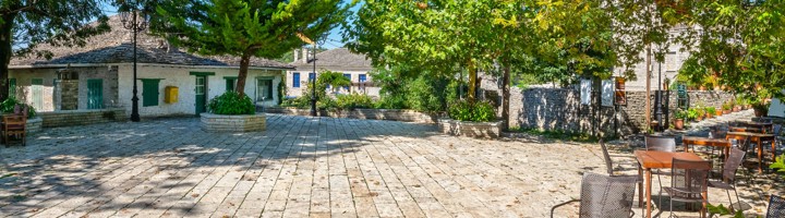 Dorpsplein van Monodendri met stenen huizen, schaduwrijke bomen en terrasstoelen in Zagori, Griekenland.
