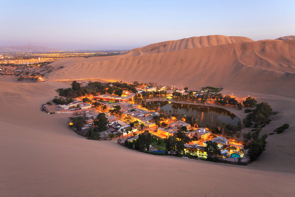 Verlichte oase Huacachina met lagune, omringd door zandduinen bij zonsondergang in Peru.
