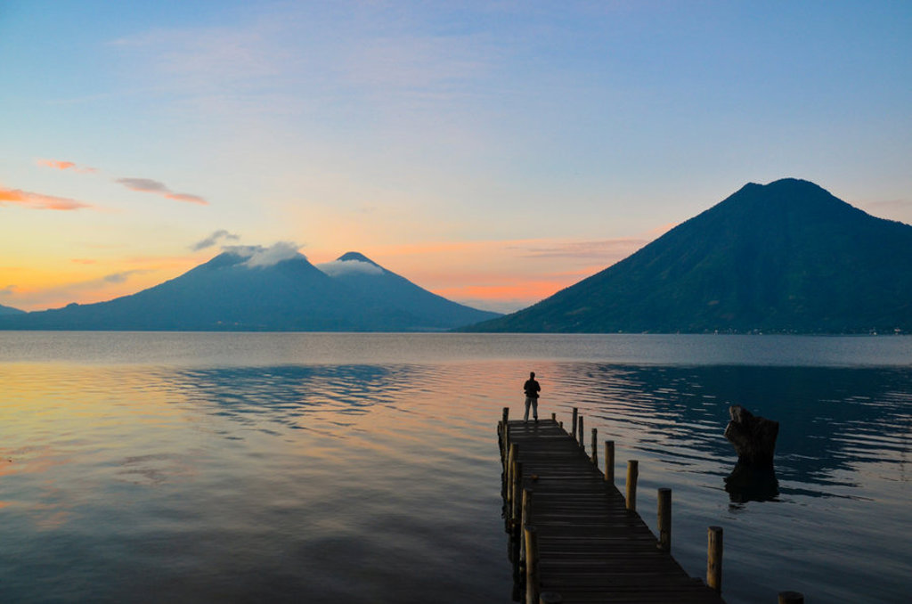 Zonsondergang bij het Meer van Atitlán in Guatemala, met vulkanen op de achtergrond en een persoon op een steiger.