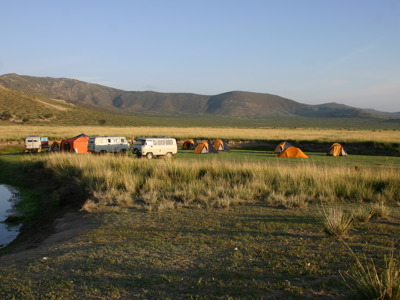 Kampeerplek met tenten en busjes in de Mongoolse steppe, met bergen op de achtergrond.