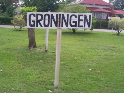 Straatbord met de tekst Groningen in Suriname, met groen gras en huizen op de achtergrond.