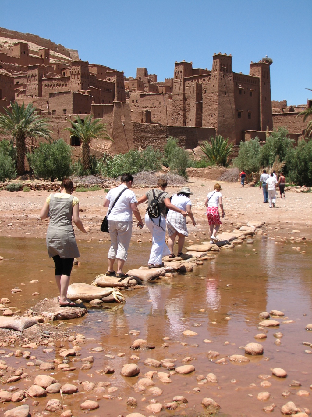 Reizigers steken via stapstenen een rivier over naar de kasbah van Aït Ben Haddou tijdens een groepsrondreis Marokko.