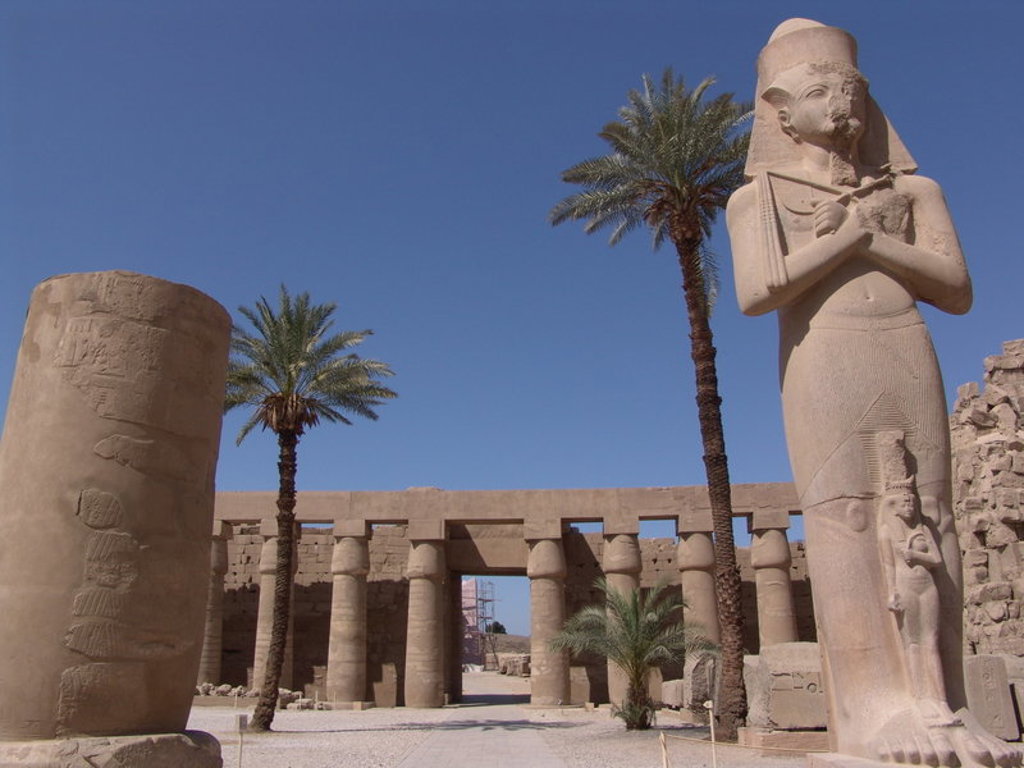 Tempelcomplex langs de Nijl in Egypte met faraobeeld, zuilen en palmbomen onder een heldere blauwe lucht.