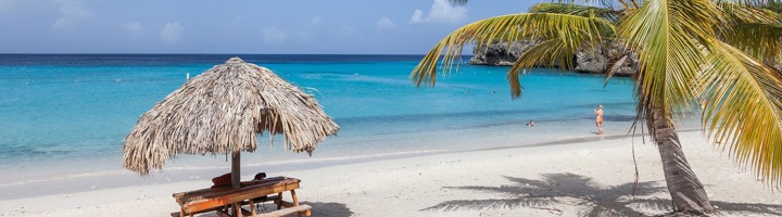 Strand op Curaçao met palmbomen, rieten parasol en helderblauw zeewater.