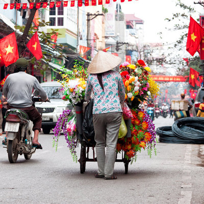 Bloemenverkoper met handkar in een drukke straat in Hanoi, Vietnam, met Vietnamese vlaggen.
