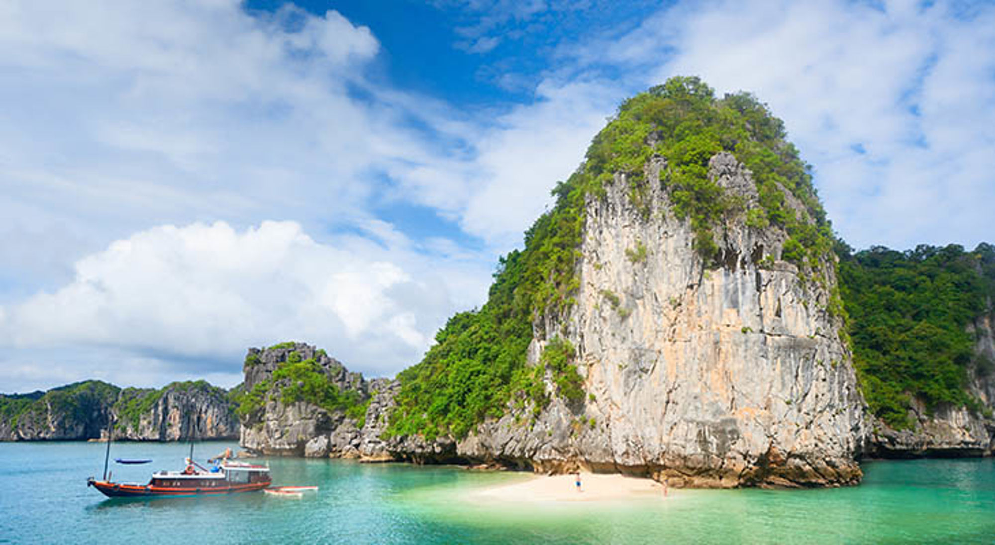 Karstrotsen en strand in Halong Bay, Vietnam, met boot op helder turquoise water.