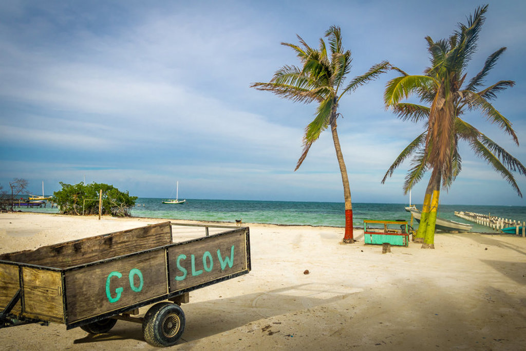 Strand in Belize met palmbomen, vissersbootjes en kar met tekst ‘Go slow’ tijdens familiereis Guatemala en Belize.
