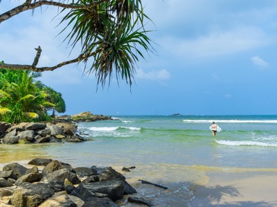 Tropisch strand in Sri Lanka met palmbomen, rotsen en een surfer die de zee in loopt.