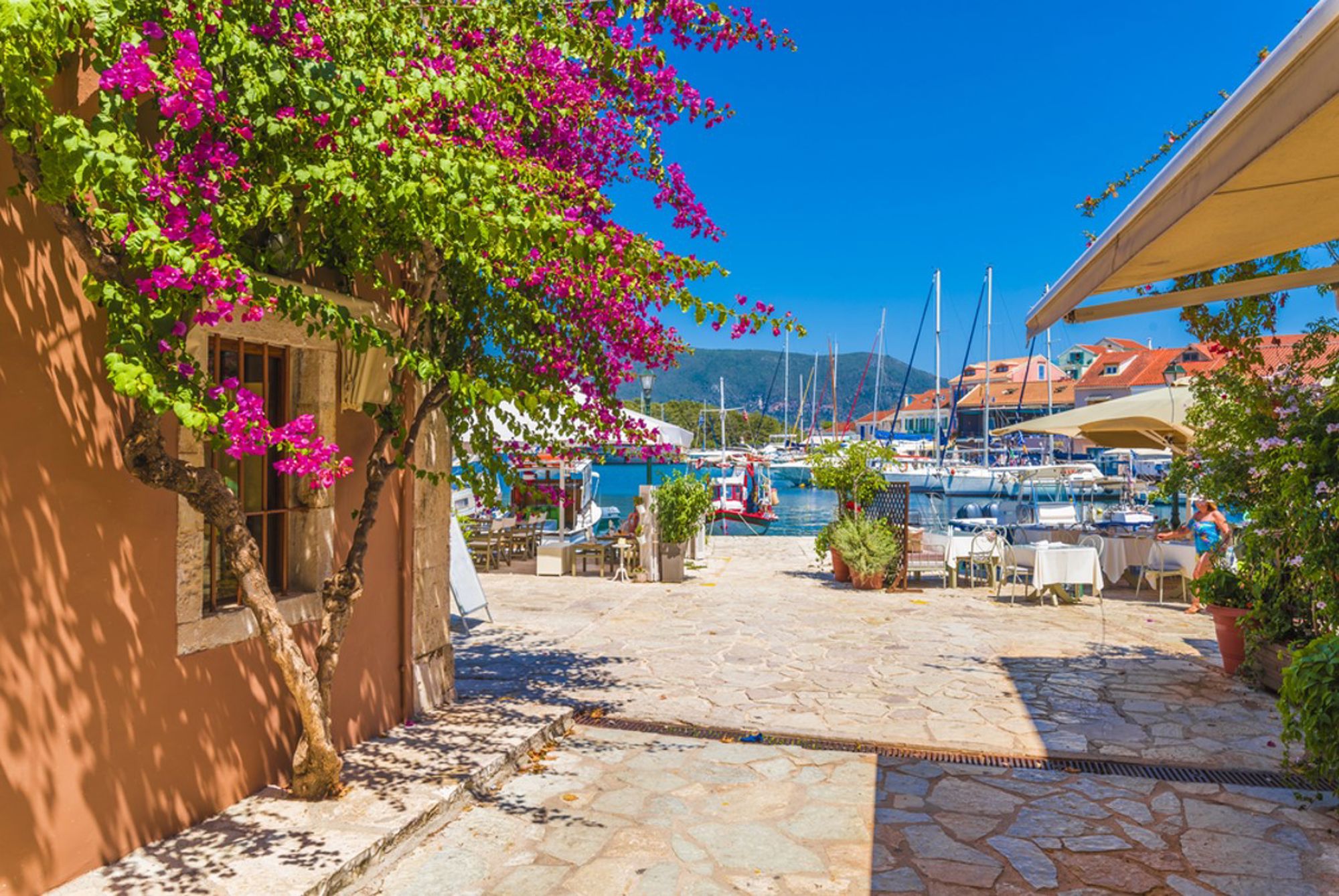 Haven in Griekenland met zeilboten, terrasjes en bougainvillea langs een zonnige promenade.