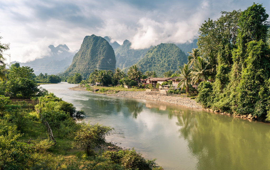 Rivierlandschap in Laos met groene karstbergen, palmbomen en dorpje langs de oever.