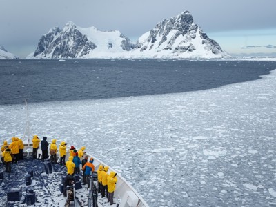 Expeditiecruise in Antarctica langs pakijs, met reizigers op het dek en besneeuwde bergtoppen op de achtergrond.