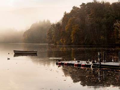 Mistig loch in Schotland met roeiboot en steiger aan de oever, omringd door herfstbos.