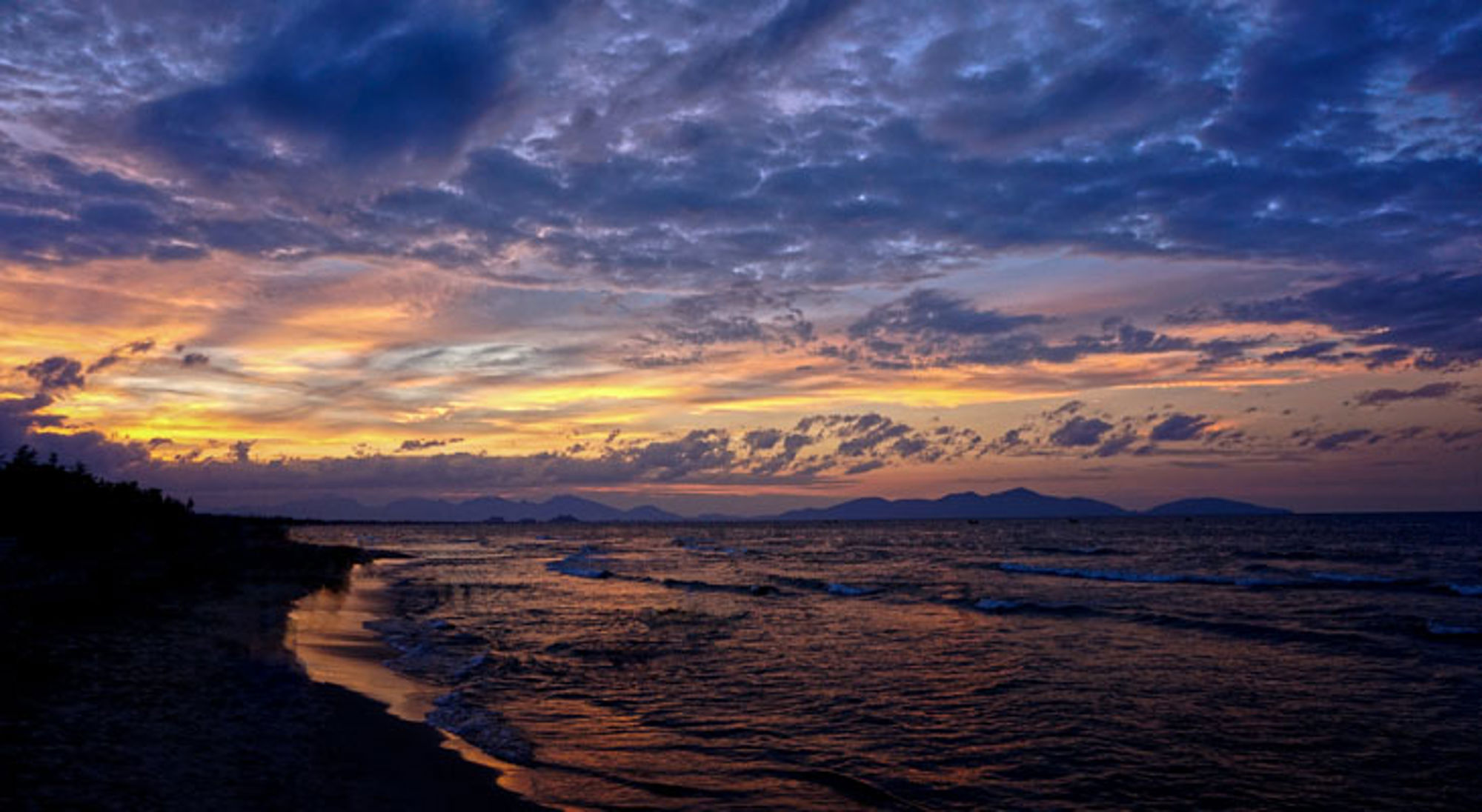 Zonsondergang boven de kust in Vietnam, met golven op het strand en donkere wolken aan de horizon.