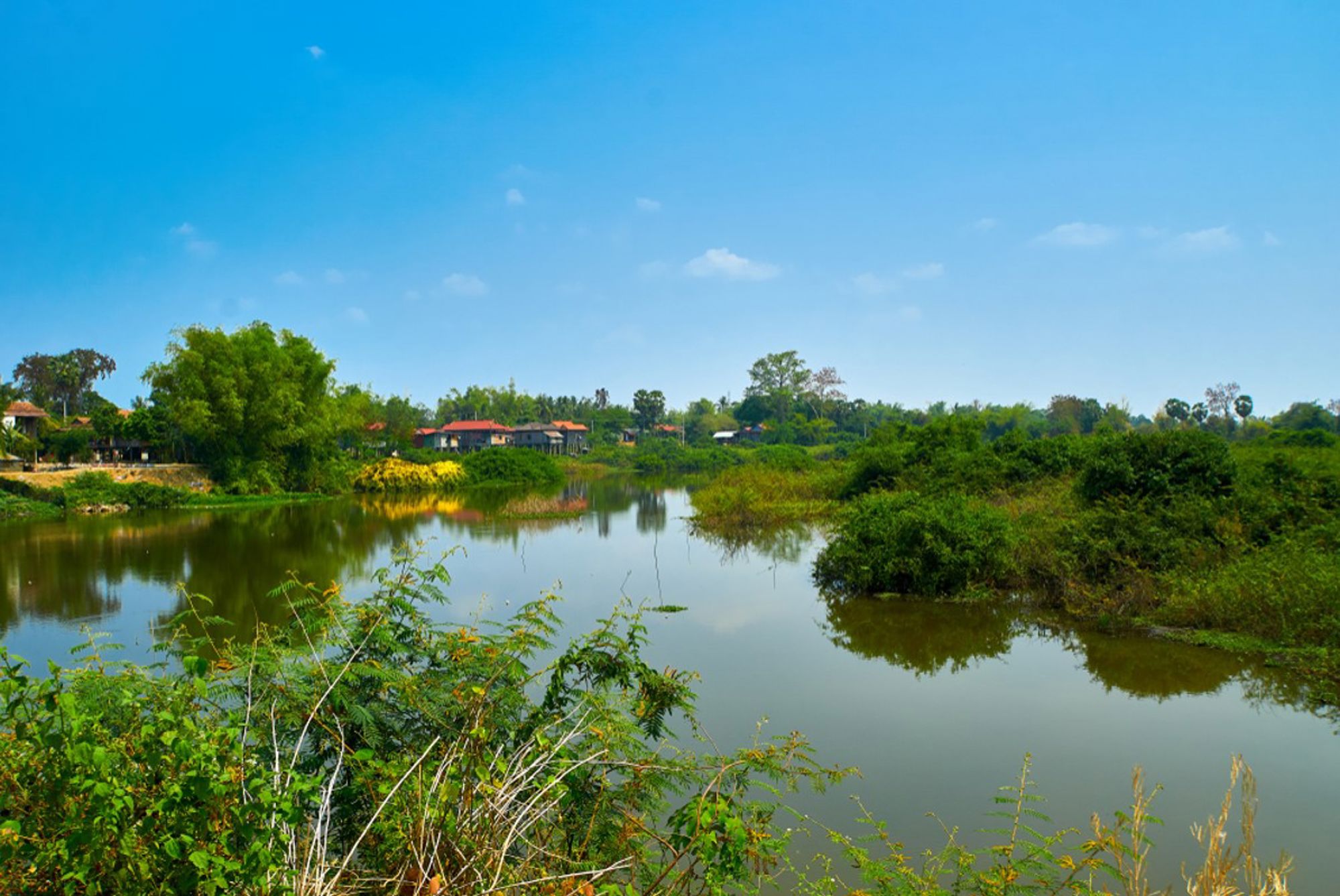 Rivierlandschap in Cambodja met groene oevers, weerspiegeling in het water en huizen aan de horizon.