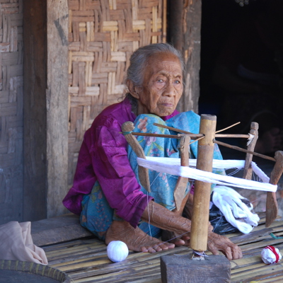 Vrouw in Pringgasela spint wit katoen op een traditionele houten spinmachine bij een huis.