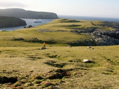 Uitzicht over ruige kust en groene heuvels met expeditieschip voor anker tijdens de route Aberdeen naar Spitsbergen.