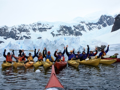 Kajakkers in Antarctica peddelen langs een gletsjer en zwaaien naar de camera.