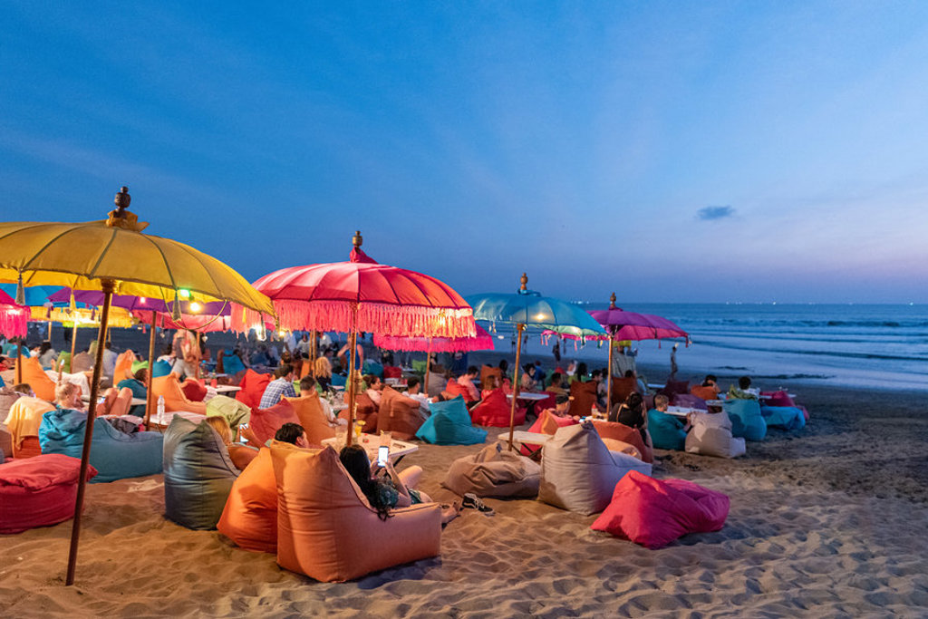 Gezellige strandbar op Bali bij zonsondergang met kleurrijke parasols en zitzakken aan zee.