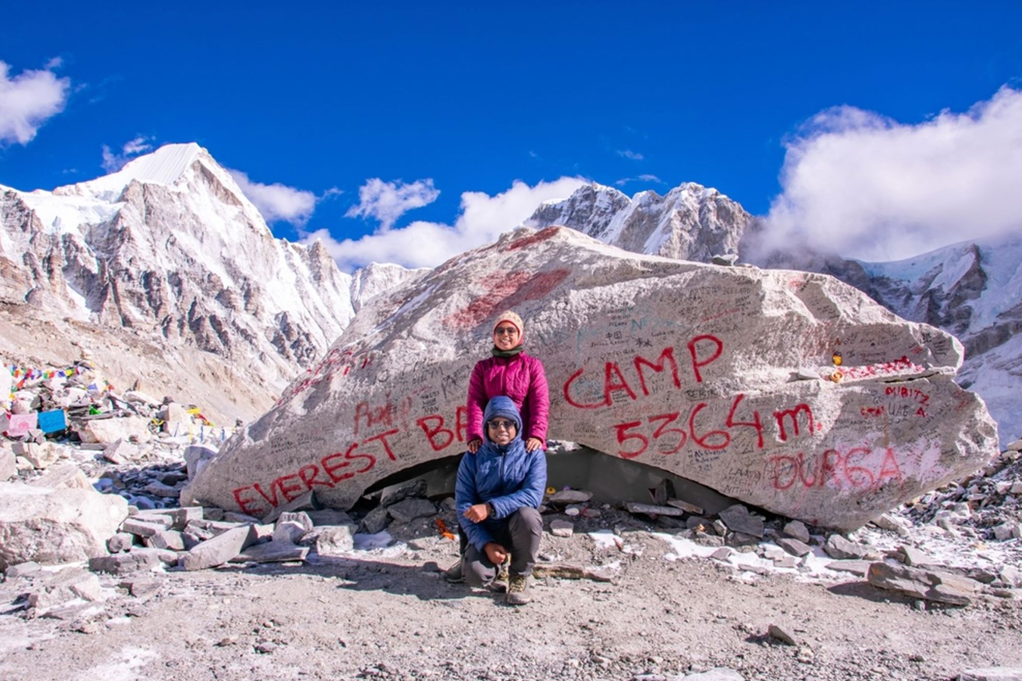 Twee wandelaars bij de steen met “Everest Base Camp 5364 m”, met besneeuwde Himalaya-toppen op de achtergrond.