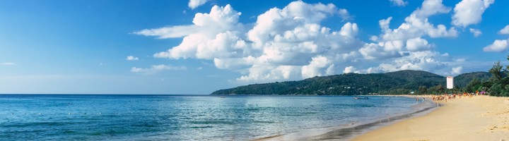 Zandstrand en rustige zee bij Karon Beach op Phuket, Thailand, met heuvels en wolkenlucht.