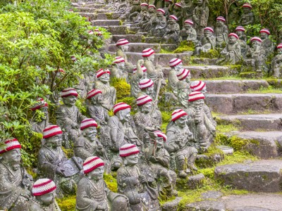 Rijen Jizo-beeldjes met rood-witte mutsjes langs mosrijke trappen bij een Japanse tempel.